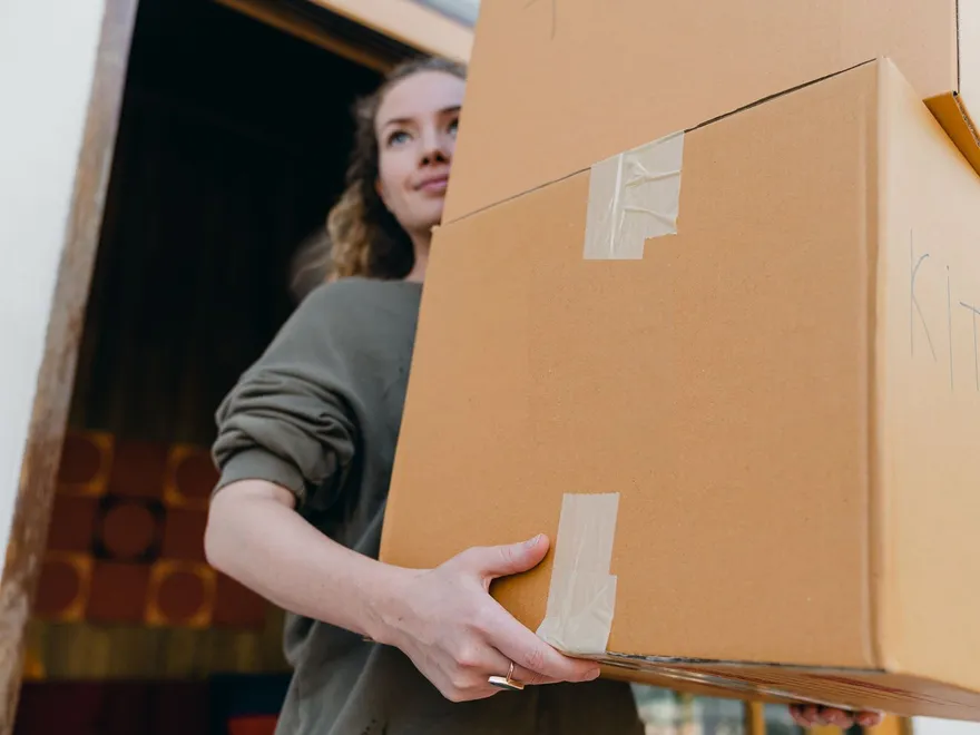A woman carrying two small moving boxes through a front door