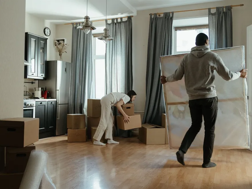 A couple carrying moving boxes and a bubble-wrapped picture frame through their lounge